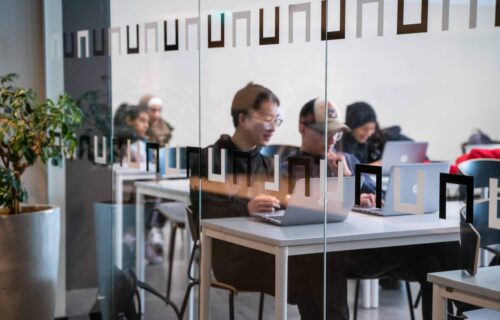 Elever i klassrum som jobbar på sina datorer. Sitter bakom en glasvägg